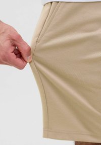 Shorts beiges avec une texture lisse et une poche latérale. Une main tire sur le tissu, montrant sa flexibilité et son élasticité.