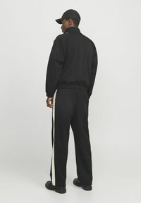 Sort træningsdragt med en jakke, der kan lynes op, og elastisk talje. Kontrasterende hvide sidelister på bukserne. Letvægtsstof, minimalistisk design.