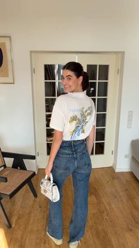 Mujer joven sonriendo por encima del hombro, vestida con una camiseta blanca con estampado floral, jeans azules de talle alto, sujetando un bolso pequeño con flores, de pie en interiores.