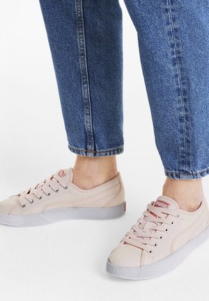 Osoba nosząca jasnoróżowe trampki Puma z białymi podeszwami w połączeniu z niebieskimi dżinsami, stojąca na białej powierzchni.