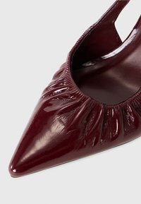 Ballerine plate bordeaux brillante à bout pointu avec un bord élastique froncé autour de l’ouverture, présentant une texture lisse et brillante en cuir verni.