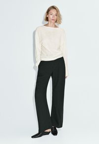 Vrouw die met haar hand in haar zak staat, gekleed in een losse witte blouse met lange mouwen, een ruimvallende zwarte broek en zwarte puntige schoenen, tegen een lichte achtergrond.