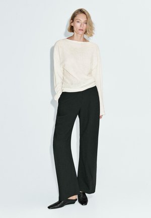 Donna in piedi con la mano in tasca, che indossa un ampio top bianco a maniche lunghe, pantaloni neri svasati e scarpe nere a punta su uno sfondo chiaro.