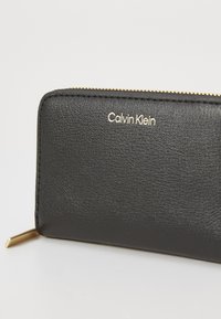 Zwarte leren portemonnee met een gestructureerde afwerking, gouden hardware-accenten en een reliëf "Calvin Klein" logo aan de voorkant. Ritsluiting zichtbaar.