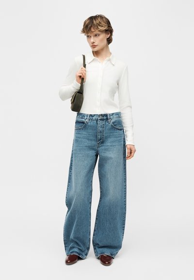 Γυναίκα που φοράει λευκό πουκάμισο με κουμπιά, φαρδιά μπλε τζιν παντελόνια, μπορντό παπούτσια και κρατά μια μικρή σκούρα τσάντα ώμου.