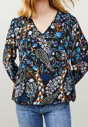 Femme portant un chemisier noir à manches longues avec un col en V, orné d’un motif coloré de fleurs et de feuilles, associé à un jean bleu.