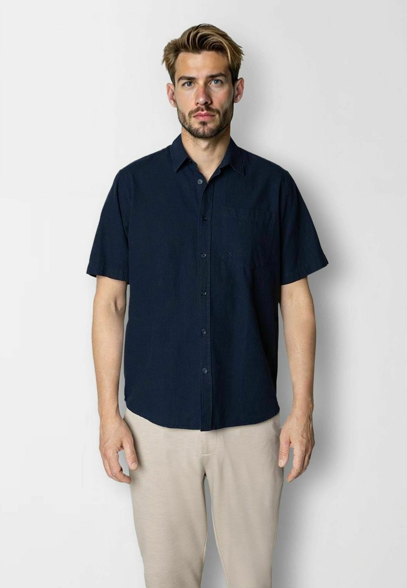 Chemise à manches courtes en tissu bleu marine foncé, avec une poche poitrine, un ourlet droit et une coupe décontractée. Associée à un pantalon beige clair.