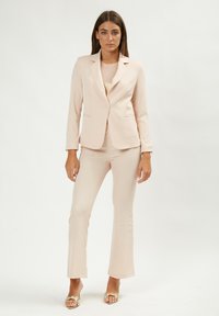 Lichtroest pak gemaakt van gladde stof, met een getailleerde blazer met één knoop, slim fit broek en open teen schoenen met gouden accenten.