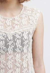 Top in pizzo con ricami floreali color avorio su uno sfondo trasparente, con un pannello grigio solido al centro e scollatura rotonda.
