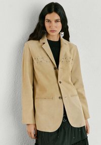 Kvinde med langt, mørkt hår iført beige blazer med knapper og perforeret design, sort top og sølvfarvede hængende øreringe foran en ensfarvet lys væg.