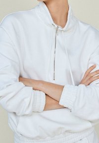 Persona che indossa un pullover bianco con mezza zip e polsini elastici, braccia incrociate sul petto.