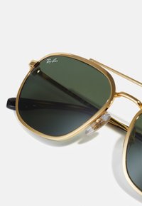 Occhiali da sole con montatura in metallo dorato e lenti verdi scure, caratterizzati da un design geometrico, morbidi naselli trasparenti e logo Ray-Ban sulla lente.