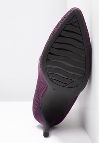 Chaussure à talon haut pointue violette, inclinée pour montrer la semelle texturée noire et le talon aiguille sur fond et surface blancs.