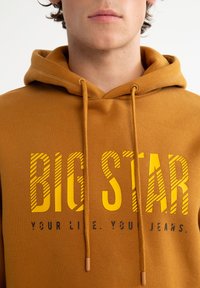 Bronzová mikina s kapucí a stahovacími šňůrkami, s výrazným žlutým proužkovaným textem "BIG STAR" a černými akcenty. Měkká, strukturovaná tkanina v uvolněném střihu.