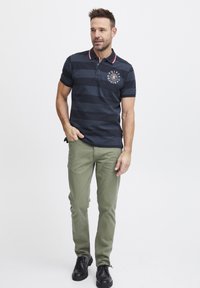 Námornícky pruhované polo tričko s červenými a bielymi detailmi na golieri, logo na hrudi, v kombinácii s olivovozelenými nohavicami a čiernymi topánkami.