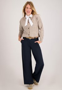 Beige vest met ronde hals, houten knopen, gecombineerd met marineblauwe wijde broekspijpen en beige sneakers, afgezet met een wit lint.