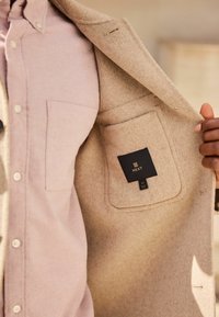 Palton bej din lână, cu o cămașă roz deschis dedesubt. Paltonul are o buzunar și o etichetă cu inscripția "NEXT" vizibilă în interior.