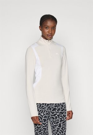 Light beige athletic top with high collar and half zip, featuring white side panels. Paired with black and white patterned leggings.