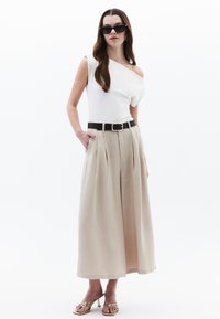 Top bianco a spalla unica abbinato a pantaloni beige a pieghe a gamba larga, accentuato da una cintura nera. La modella indossa occhiali da sole neri e sandali dorati.