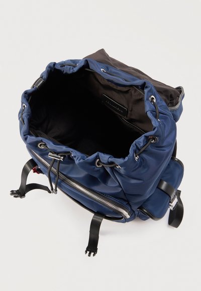 Sac à dos bleu marine à cordon avec intérieur noir, poche zippée avant, bretelles réglables et œillets argentés sur fond blanc.