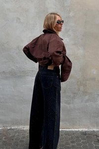 Rjava suede kratka jakna s ohlapnimi črnimi denim hlačami z diskretnimi pikčastimi vzorci in oznako NA-KD na pasu. Črna sončna očala.