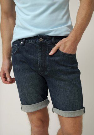 Hombre con pantalones cortos de mezclilla azul oscuro remangados con bolsillos delanteros y una camiseta azul claro, mano en el bolsillo.