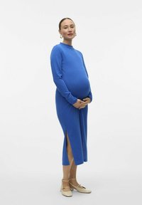 Mėlyna banguota nėščiųjų suknelė su ilgomis rankovėmis, šoninėmis skeltuvėmis ir laisvu siluetu, derinama su šviesios spalvos slingback batais.