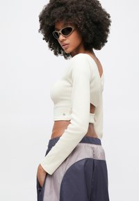 Γυναίκα με σγο υρά μαλλιά που φοράει μπεζ crop top με μακριά μανίκια και ανοιχτή πλάτη, παντελόνι δίχρωμο σε μπλε ναυτικό και γκρι, και μικρά μπεζ γυαλιά ηλίου, ποζάροντας πλευρικά.