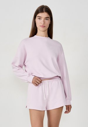 Junge Frau mit langen braunen Haaren, die einen hellrosa Sweatshirt und passende Bundfalten-Shorts trägt, steht vor einem schlichten hellen Hintergrund.