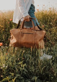 Braune Leder-Reisetasche mit doppelten Griffen, einem Schultergurt und einer vorderen Reißverschlusstasche, vor einem Hintergrund aus Gras und wilden Blumen.