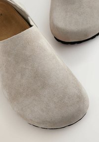 Sabots en suède gris clair avec une forme arrondie et une texture lisse. La semelle est foncée et texturée pour une meilleure adhérence. Les éléments de design sont minimalistes.