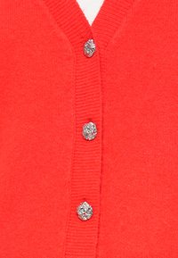 Cardigã vermelho em tricô com botões prateados texturizados e uma peça interior branca parcialmente visível na parte superior.