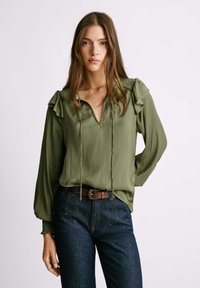 Olive green blouse with ruffled shoulders, V-neckline, and decorative buttons. Paired with dark blue jeans and a brown belt with a circular buckle.