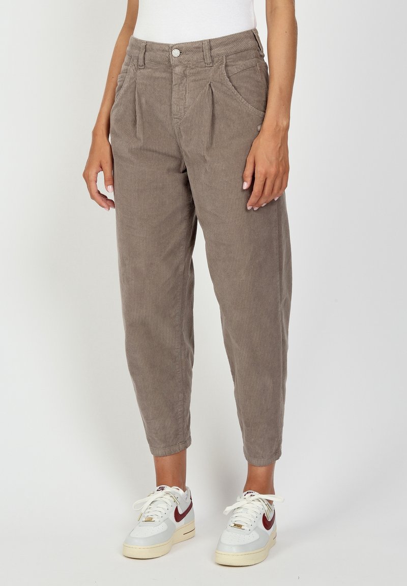 Personne portant un pantalon en velours côtelé taupe taille haute avec des plis et des baskets blanches avec un swoosh rouge, debout devant un fond blanc.