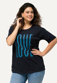 Camiseta negra de tejido suave con un gran gráfico en texto azul que dice "AMOR". Cuello redondo y mangas cortas, combinada con jeans de color azul claro.