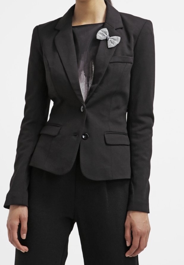 Femme portant un blazer noir ajusté à deux boutons avec un nœud papillon rayé noir et blanc sur le revers gauche.