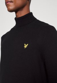 Maglione a collo alto nero in materiale morbido, con un piccolo logo di un uccellino giallo ricamato sul lato sinistro del petto.
