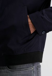 Hand gedeeltelijk in de zak van een marineblauwe jas met een dichtgeknoopte manchet, gedragen over grijze denim jeans.