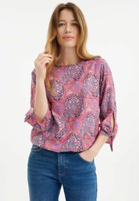 Multifarbige Paisleymuster-Bluse in Pink, Lila und Blau, mit Rundhalsausschnitt und Dreiviertelärmeln mit Bindebändern. Tragen mit blauen Jeans.