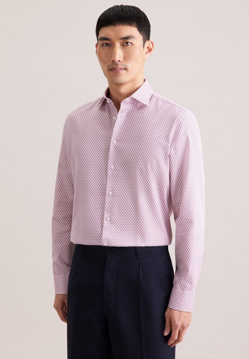 Homme portant une chemise à manches longues avec un motif géométrique rose et un pantalon sombre, debout devant un fond clair uni.