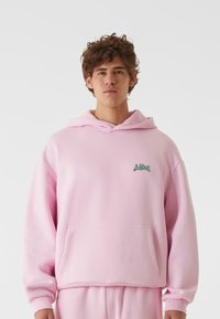 Haut à capuche rose clair en tissu doux, comprenant une poche avant et un logo vert sur la poitrine. Le design comprend une coupe décontractée et une capuche avec cordon.