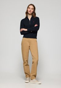 Sweatshirt zippé navy avec un ourlet et un col côtelés, associé à un chino beige et des baskets blanches, se tenant devant un fond neutre.