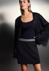 Robe noire à la silhouette ajustée, associée à un cardigan noir texturé et duveteux. La tenue présente une ceinture en argent étincelant à la taille.