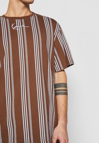 Muž v hnědo-modrém tričku s vertikálními pruhy a podpisovým detailem u límce, s černým tetováním ve třech pruzích na předloktí.