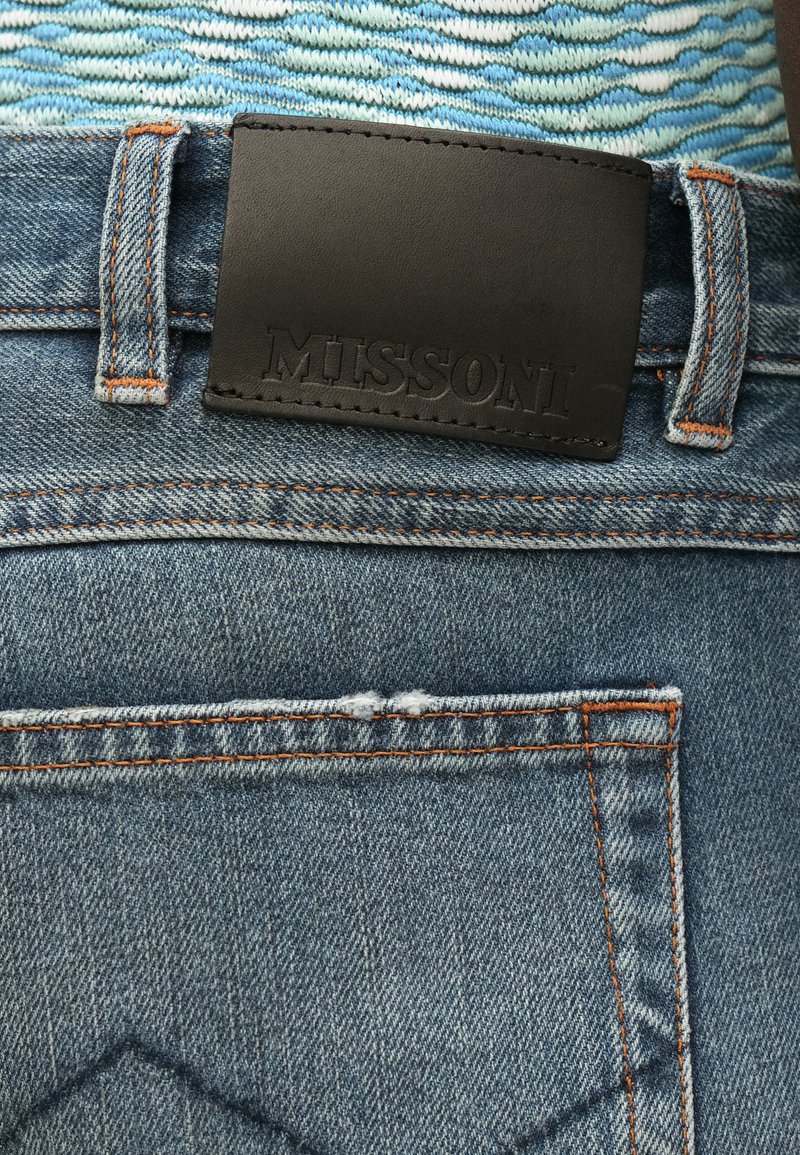 Jean en denim avec un patch en cuir noir portant l'inscription "MISSONI". Il présente un tissu bleu classique, des coutures orange et des détails visibles sur la poche.
