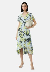 Blumenkleid in Hellblau, mit gelben und schwarzen Mustern, ausgestelltem Saum, kurzen Ärmeln, kombiniert mit einer kleinen weißen Handtasche und Sandalen.