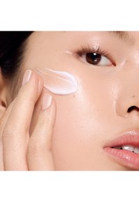 Moisturizing cream applied to light, smooth skin; the cream is a soft, opaque white, shown on a fingertip and cheek for texture.