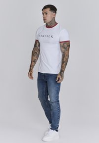 Korte mouwen witte T-shirt met rode rand en zwart logo, gecombineerd met blauwe denim jeans en witte sneakers. Model staat naar links gericht.