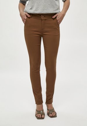Femme portant un pantalon marron près du corps et des sandales ouvertes vert olive, debout les mains dans les poches devant un fond uni.