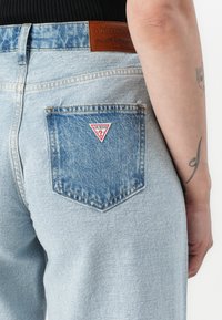 Light blue denim jeans featuring a triangular red and white logo patch on the back pocket. The fabric has a faded, soft texture.
